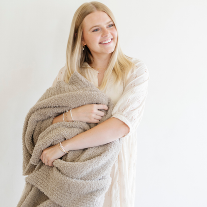 A woman smiles and holds a Bamboni® blanket by Saranoni. This ultra-soft blanket is a taupe blanket and made from a premium blend of bamboo rayon and poly microfiber, offering a lightweight, breathable, and fluffy feel. Perfect as a throw blanket, luxury blanket, plush throw, or soft blanket, it adds warmth and elegance to any living room, bedroom, or home décor setting. Designed for ultimate comfort, this buttery-soft Saranoni blanket is ideal for gifting. Enjoy this cozy blanket for years to c