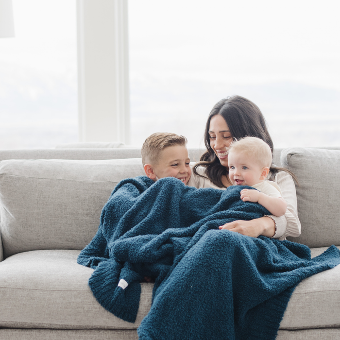 A mother and her kids sit with a Bamboni® blanket by Saranoni. This ultra-soft blanket is a blue blanket and made from a premium blend of bamboo rayon and poly microfiber, offering a lightweight, breathable, and fluffy feel. Perfect as a throw blanket, luxury blanket, plush throw, or soft blanket, it adds warmth and elegance to any living room, bedroom, or home décor setting. Designed for ultimate comfort, this buttery-soft Saranoni blanket is ideal for gifting. Enjoy this cozy blanket for years