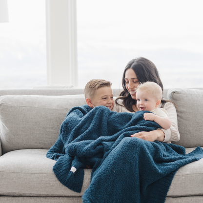 A mother and her kids sit with a Bamboni® blanket by Saranoni. This ultra-soft blanket is a blue blanket and made from a premium blend of bamboo rayon and poly microfiber, offering a lightweight, breathable, and fluffy feel. Perfect as a throw blanket, luxury blanket, plush throw, or soft blanket, it adds warmth and elegance to any living room, bedroom, or home décor setting. Designed for ultimate comfort, this buttery-soft Saranoni blanket is ideal for gifting. Enjoy this cozy blanket for years