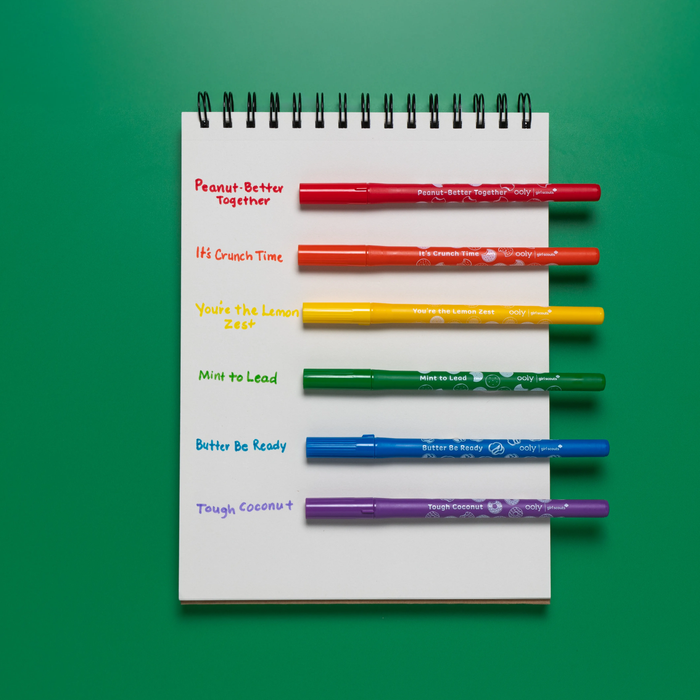 Sketchbook with OOLY x Girl Scouts Cookie Scented Markers resting next to handwritten scent names as swatches