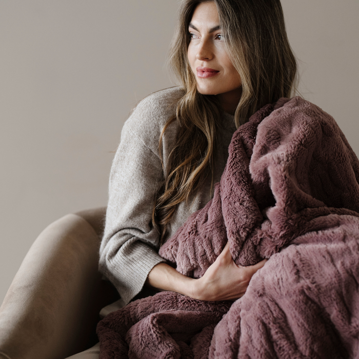 A woman sits in a chair and is holding a dusty mauve patterned faux fur blanket. This soft blanket is a luxury blanket and a Saranoni blanket. The luxurious texture of this blanket makes it perfect for gifting, relaxing, or using for home decor.