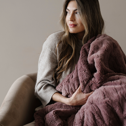 A woman sits in a chair and is holding a dusty mauve patterned faux fur blanket. This soft blanket is a luxury blanket and a Saranoni blanket. The luxurious texture of this blanket makes it perfect for gifting, relaxing, or using for home decor.