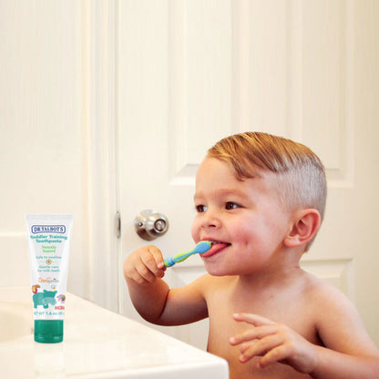 A young child with short blond hair smiles while brushing their teeth with Dr. Talbot's Toddler Toothbrush and Toothpaste Set by the bathroom sink. The non-toxic, fluoride-free toothpaste is visible in the foreground.