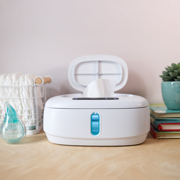 Dr. Talbot's Touch-Free Wipe Warmer dispenses a baby wipe from its open lid on a wooden surface, surrounded by rolled diapers in a basket, books, a nasal aspirator, and a potted succulent.